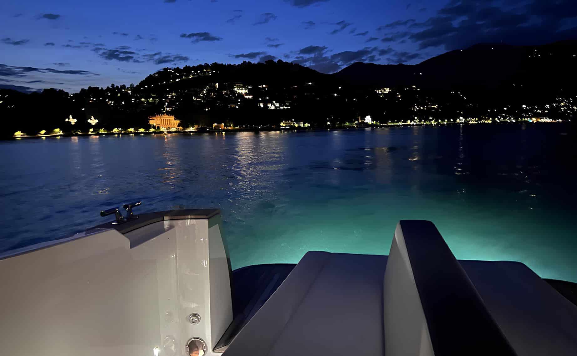 Poppa del Limotender con luci subacquee verdi e costa del Lago di Como illuminata durante crociera notturna di lusso.