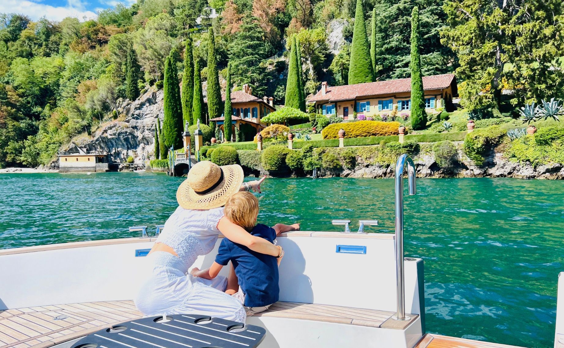 Mother and son on luxury boat pointing at elegant lakeside villa surrounded by tall cypress trees.
