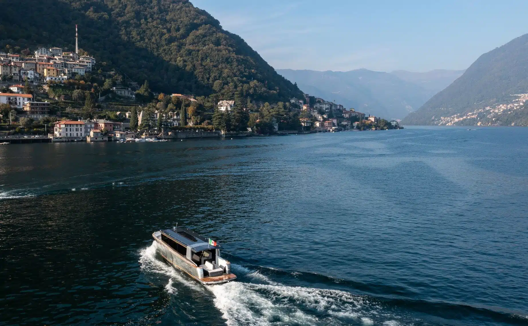 Drone view of Limotender underway near lakeside villages, leaving a curved white wake.